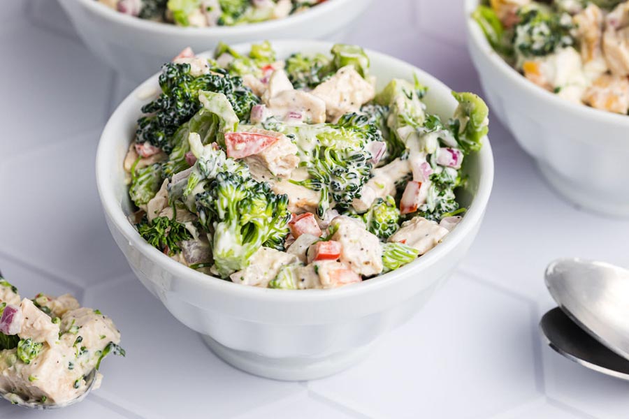 A small bowl of broccoli salad coated in a creamy dressing with chunks of chicken and red pepper.