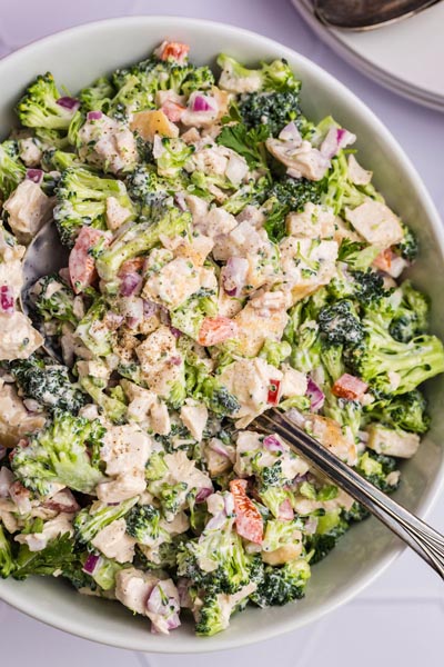 A serving spoon digging into a big bowl of creamy broccoli chicken salad.