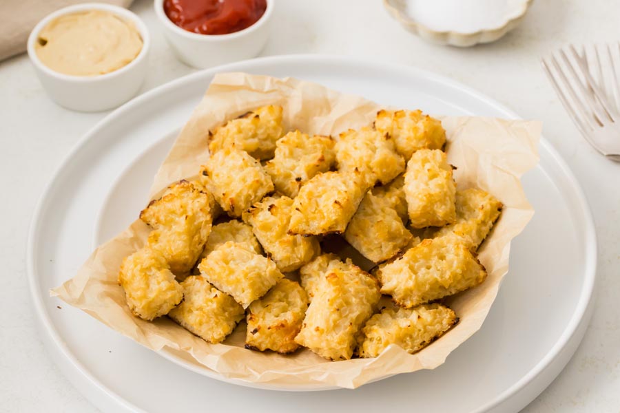 A bowl of cheesy cauliflower tots on a plate with mustard and ketchup in the background.