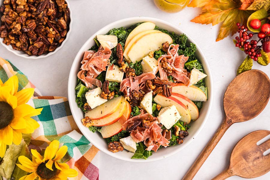 A bowl of kale salad with harvest fruits and veggies including apple, kale, prosciutto and spiced nuts.