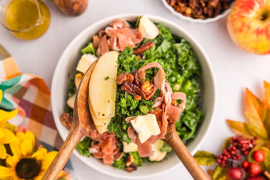 Salad tongs holding up a serving of harvest salad with nuts and apple.