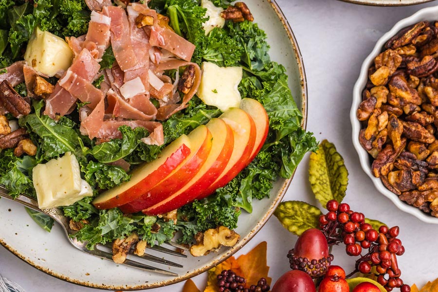 Kale salad with sliced apple, sliced prosciutto and brie cheese next to a bowl of candied nuts.