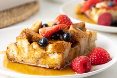 A slice of high protein french toast casserole on a plate covered with sugar-free maple syrup and topped with raspberries, blueberries and strawberries.