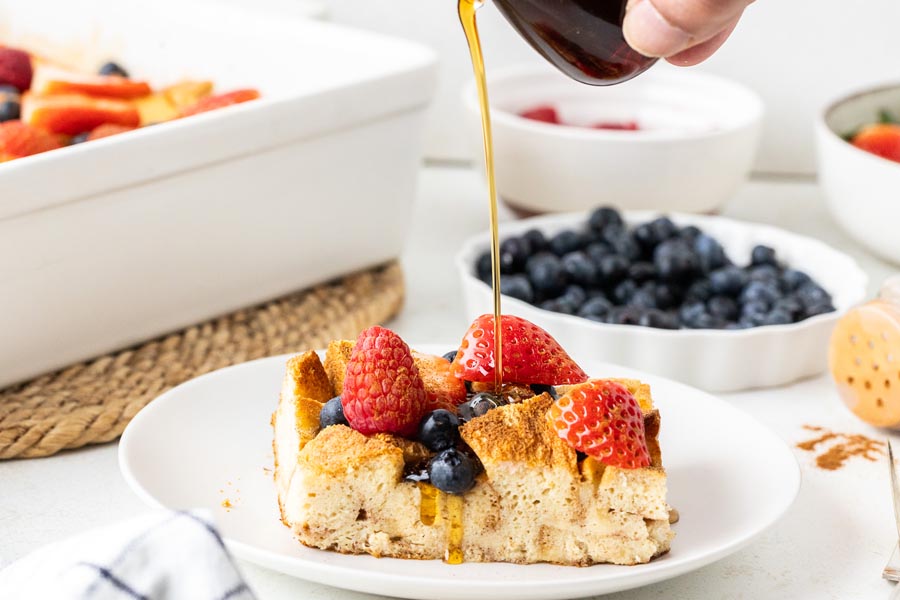 A hand pouring syrup over a slice of french toast casserole that is topped with raspberries, blueberries and strawberry slices.