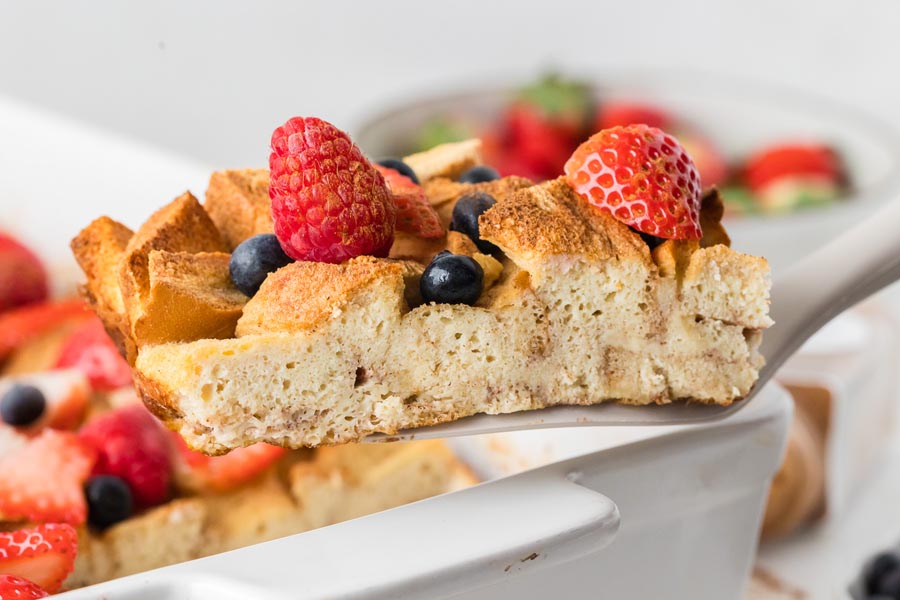 A spatula holding a slice of custardy french toast casserole topped with fresh berries.