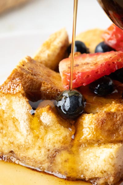 A stream of sugar free maple syrup being poured on top of a slice of french toast casserole with berries on top.