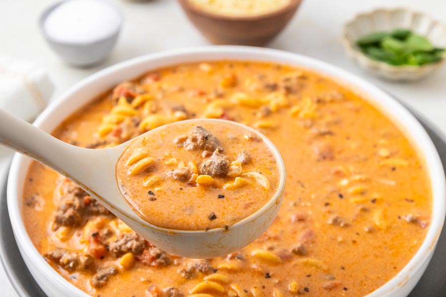 A ladle holding a scoop of tomato soup filled with noodles, ground beef and tomatoes.