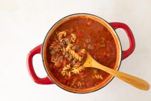 A red dutch oven filled with tomato based soup with ground beef and pasta.