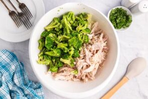 Cooked broccoli florets and shredded chicken side by side in a large white bowl.