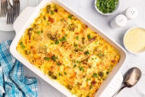 A finished chicken divan casserole on the counter topped with parsley and next to a spoon, parmesan cheese and more parsley.