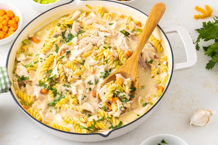 A white skillet with spiral noodles, chicken and vegetables with a wooden serving spoon inside.