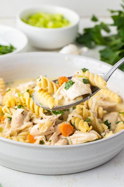 A spoon holding a bite of casserole over a bowl of spiral noodles, chicken and carrots.