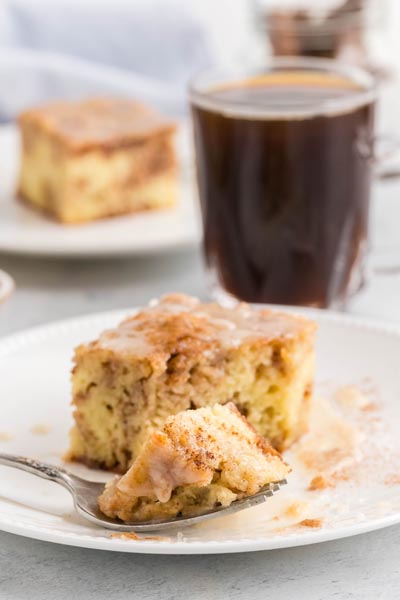 A bite of cinnamon coffee cake on a fork sitting on a plate in front of a cup of coffee.