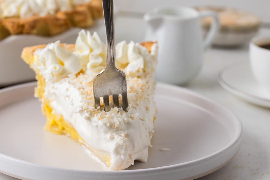 A fork digging into a slice of coconut cream pie on a plate.