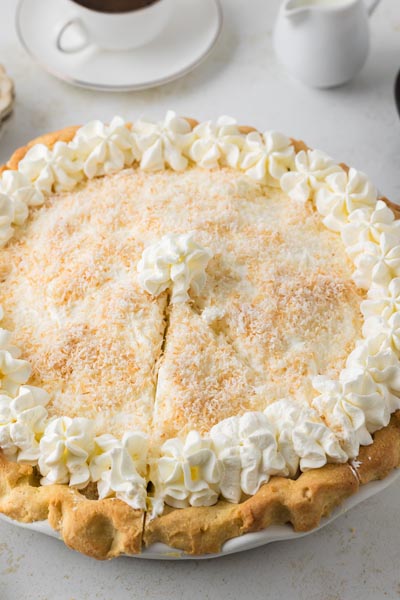 A slice carved out of a whole coconut cream pie next to a cup of coffee.