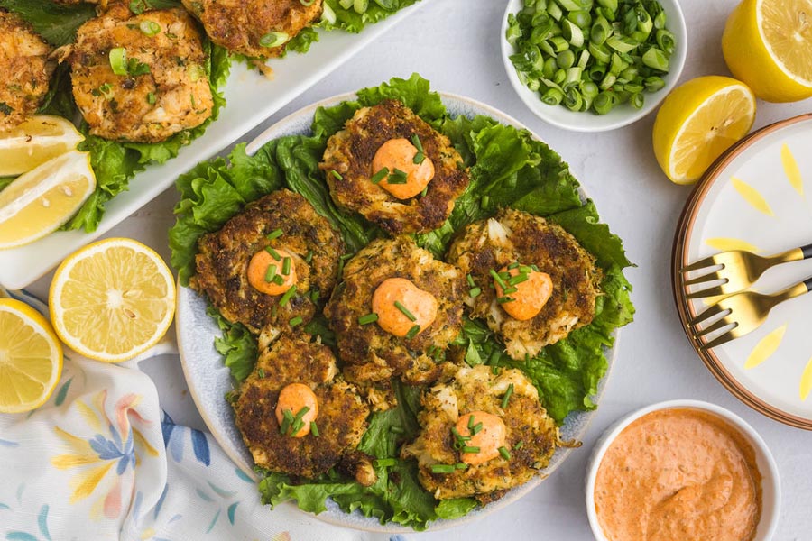A platter of crab cakes topped with sauce next to fresh lemon and green onions.