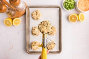A cookie scoop filled with crab cake mixture over more crab cakes on a tray.