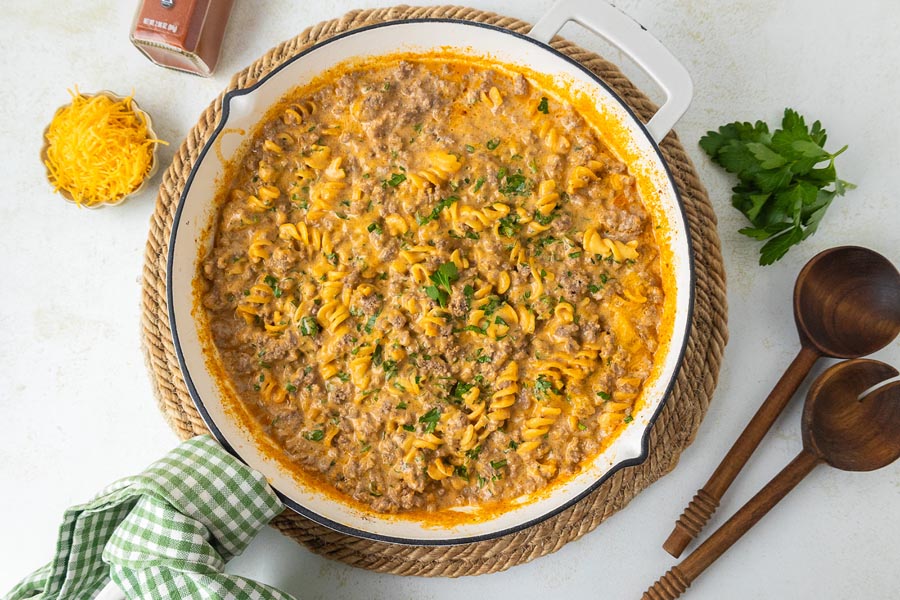 Cheesy hamburger casserole in a skillet next to serving spoons and shredded cheese.