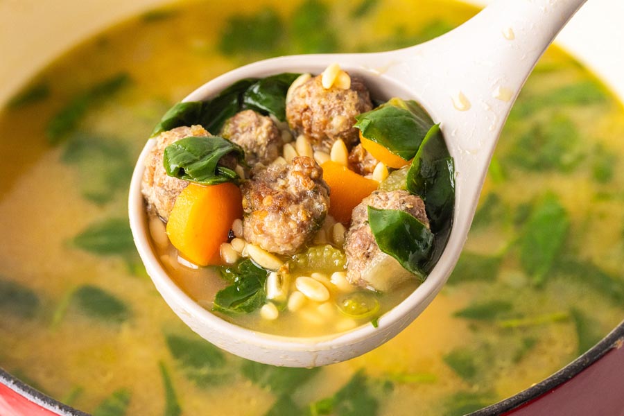 A ladle holding a serving of italian wedding soup with meatballs, spinach, carrot and rice pasta.