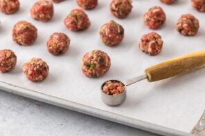 A teaspoon filled with meatball mixture on a baking sheet next to small meatballs formed on the tray.