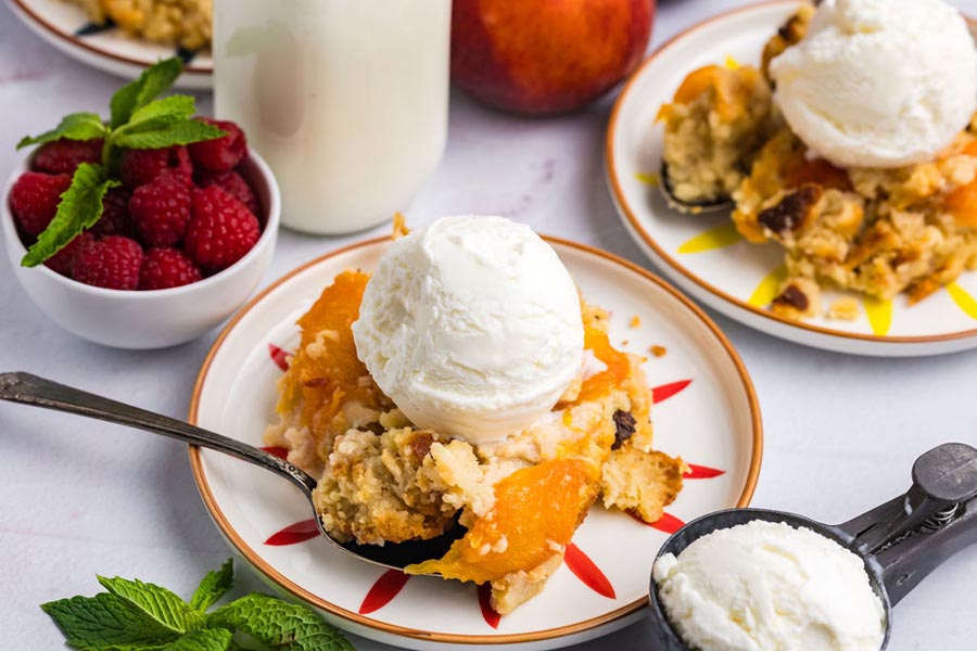 A spoon holding some peach cobbler on top of plate with fresh raspberries nearby.