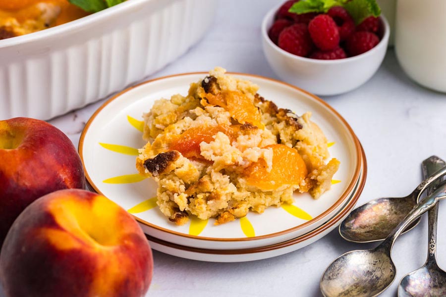 A scoop of peach cobbler on a plate next to whole peaches.