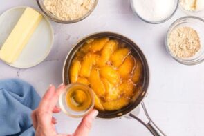 A hand holding peach flavoring over a pot full of peaches.