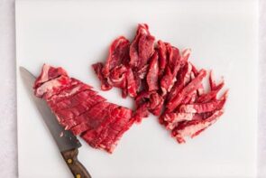 Flank steak cut into thin strips on a cutting board with a knife next to it.