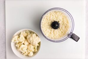 Cauliflower florets sit next to a food processor with riced cauliflower inside.