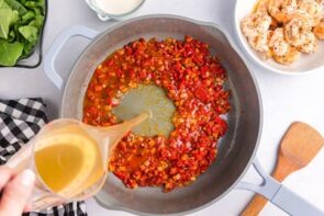 A hand pouring chicken broth into a skillet with cooked bell pepper.