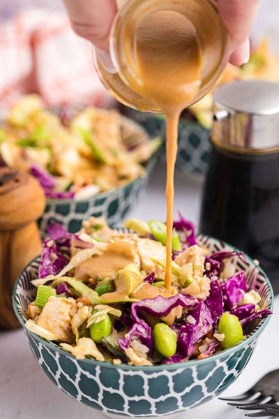 A hand pouring creamy peanut dressing on a thai chicken salad bowl.